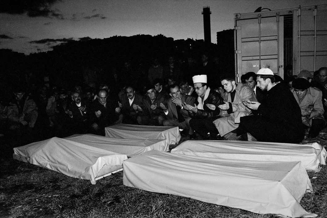 Prayers are led by a mufti over the coffins of children and adults killed by Serbian mortar fire on a school. According to muslim tradition, the dead are to be buried before sunset. Because of the dangers of sniper fire in exposed cemetaries, mourners wait until the last minutes of daylight for the rites to the dead. Sarajevo. 1993.