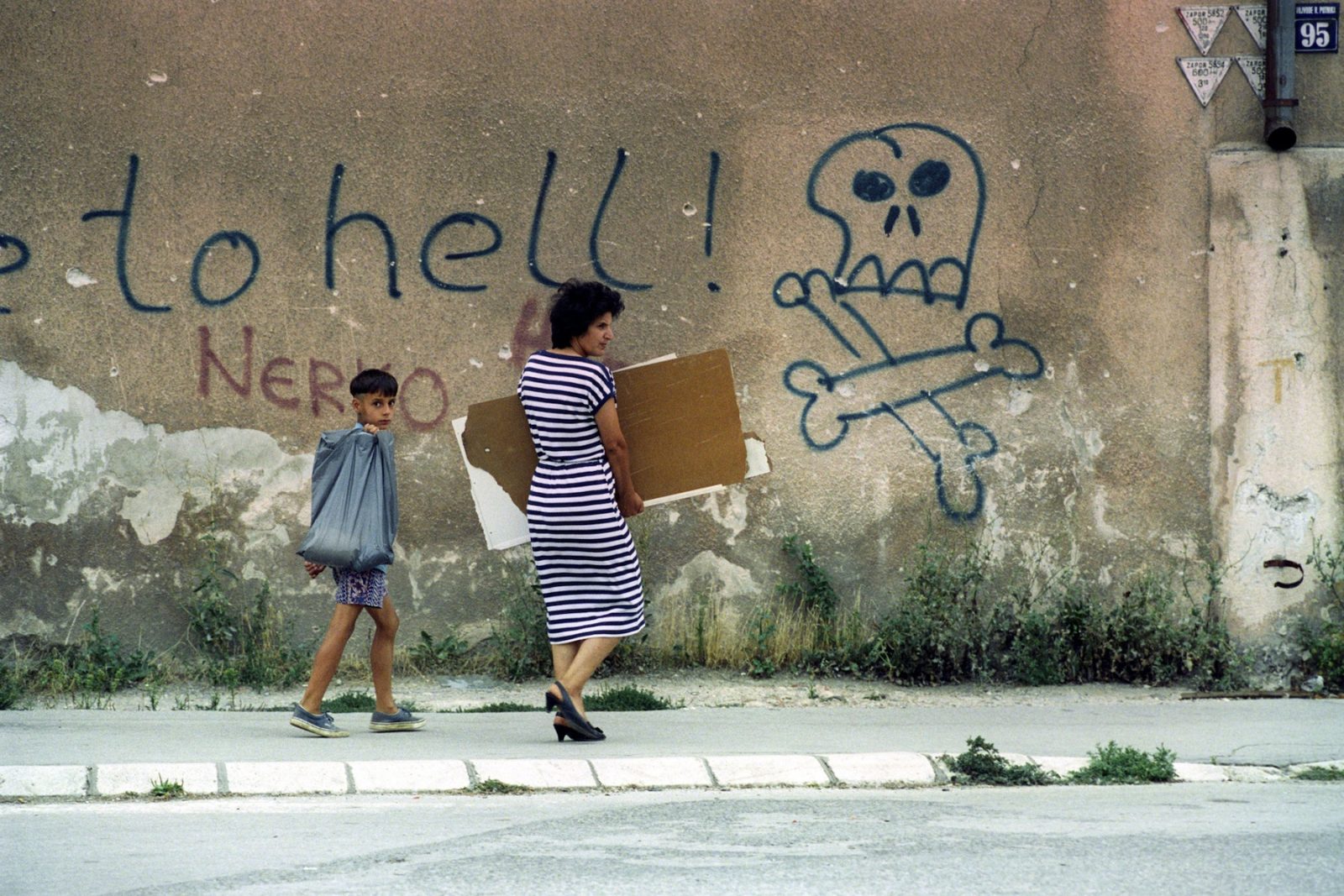 Accompanied by her son, a mother carries panels of wood along a street and past graffiti showing a skull and crossbones along with the words ' welcome to hell' in Sarajevo, Bosnia and Hercegovina, October 1993.