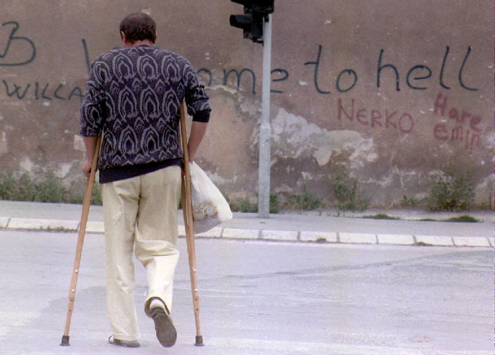 A man on crutches walks across the area of Sarajevo known as "Sniper Alley" 28 August 1993, near a wall with graffiti that reads "Welcome to Hell." The Bosnian parliament rejected 27 Aug a U.N. peace plan unless former Muslim-majority areas are joined to a proposed Muslim-dominated Bosnian republic. (Photo by GABRIEL BOUYS / AFP)