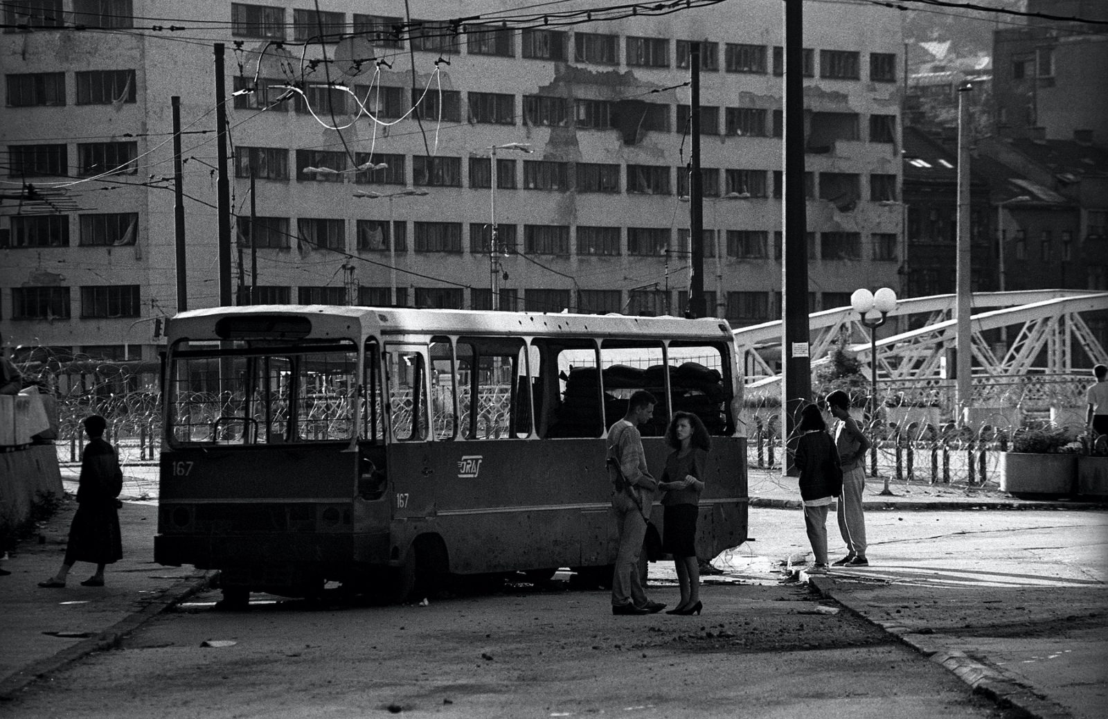 A couple chatting next to a destroyed bus near the front line with Grbavica, in the centre of Sarajevo. Sarajevo, Bosnia and Herzegovina, August 1993