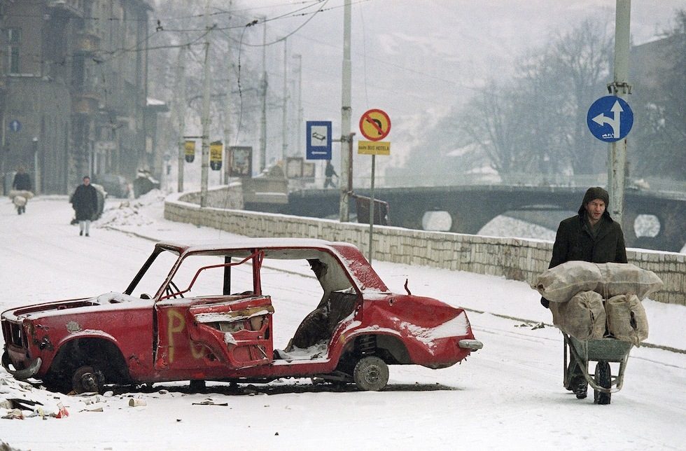 A Sarajevo man pushes his wheelbarrow full of wood, past a car that was destroyed earlier during the siege of Sarajevo on Tuesday, March 9, 1993. The street is normally in open view of Serbian snipers, but light snow or fog sometimes shields pedestrians on the city streets below from snipers. (AP Photo/Michael Stravato)