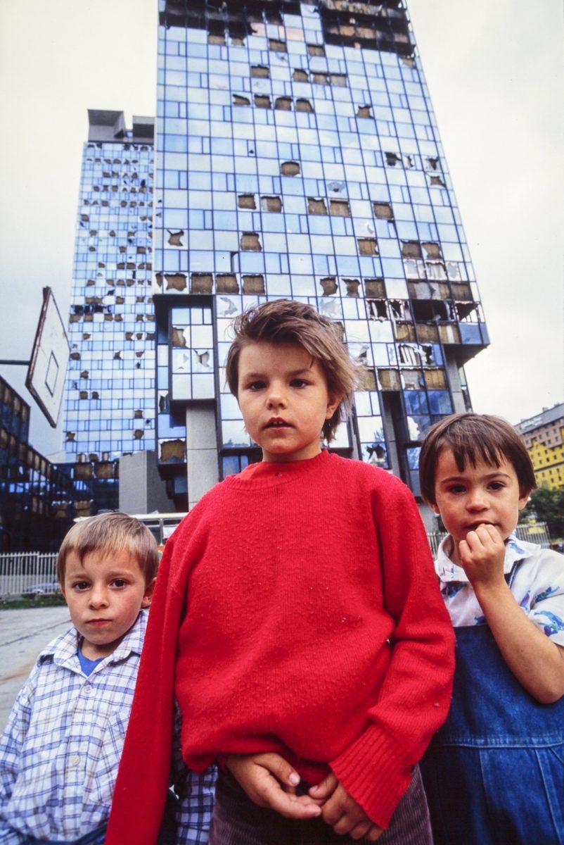 Three young Bosnian children stand near a damaged office building in war-torn Sarajevo. 13 Jul 1992, Sarajevo, Bosnia and Herzegovina