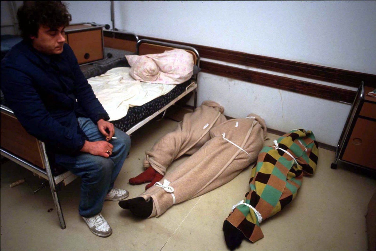 Bodies of deceased retirement home residents wrapped in blankets. Caregiver smoking a cigarette whilst waiting for bodies to be transported away. Sarajevo, Dobrinja, Decembre 1992. Photo Copyright © Peter Kullmann| Zone5 | fullframeRAW